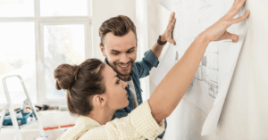 A woman and man smile while examining a blueprint on a wall. The bright room has a ladder and tools, suggesting renovation or remodeling work.